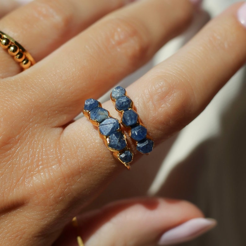 May include: Two gold rings with blue sapphire stones. The stones are faceted and have a rough, natural look. The rings are stacked on top of each other.