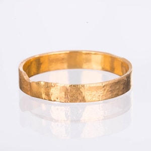 Op de afbeelding: Een goudkleurige ring met een getextureerd oppervlak. De ring heeft een gehamerde uitstraling en een eenvoudig bandontwerp. De ring wordt gepresenteerd op een wit oppervlak, dat de vorm weerspiegelt. De diameter is ongeveer 2 cm.