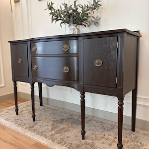 May include: A dark brown antique buffet with two cabinet doors, two drawers, and decorative brass hardware. The buffet has four slender, turned legs and is set on a patterned rug. A vase of greenery sits on top.