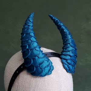 May include: A pair of teal-colored, textured horns attached to a black headband. The horns are curved and have a scale-like pattern. The headband is worn on a mannequin head against a green background.
