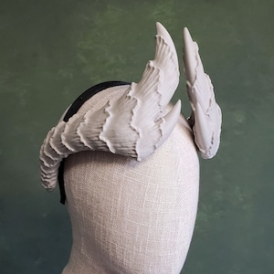 May include: A white headband with two sculpted horns, resembling a crown or headdress. The horns are made of a smooth, matte material and have a slightly textured surface.