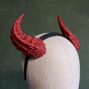 May include: Red devil horns headband with a black elastic band. The headband is on a white fabric mannequin head.