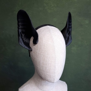 May include: Black satin bat ear headband. The ears are large and curved, attached to a thin black headband. The ears are made of a shiny, textured fabric. The headband is displayed on a mannequin head against a green background.