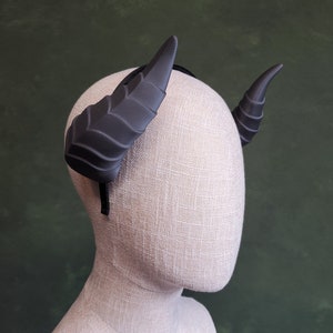 Puede incluir: Una diadema negra con dos cuernos grandes, negros y texturizados. La diadema está en una cabeza de maniquí blanca.