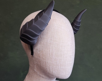 May include: A black headband with two large, black, textured horns. The headband is on a white mannequin head.