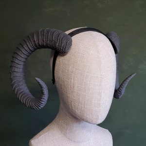 May include: A gray ram horn headband. The horns are large, curved, and textured, attached to a black headband. The horns are a dark gray color and are displayed on a mannequin head.