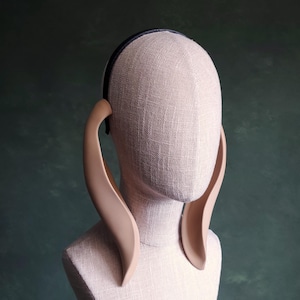 Puede incluir: Un par de grandes pendientes de oreja curvos de color beige, hechos de un material liso y flexible. Los pendientes están unidos a una diadema negra.