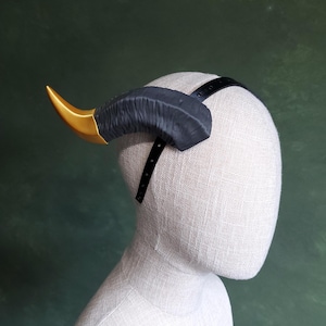Peut inclure: Un serre-tête avec une corne noire et dorée. La corne est noire avec une pointe dorée. Le serre-tête a une sangle noire qui fait le tour de la tête. Les cornes sont fixées à une bande noire qui repose sur la tête.