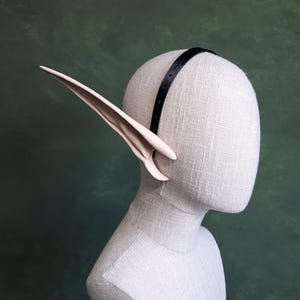 May include: A pair of pale pink elf ears attached to a black headband, displayed on a mannequin head. The ears are long and pointed, with a curved shape. The headband is thin and has small holes. The background is a solid green.
