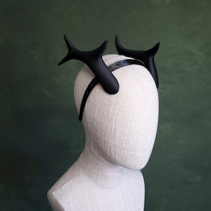 May include: A black antler headband, featuring two stylized, curved antlers. The antlers are mounted on a thin, black headband. The headpiece is displayed on a mannequin head against a green background.