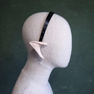 May include: A white elf ear headband with a black adjustable strap. The headband is worn on a white mannequin head.
