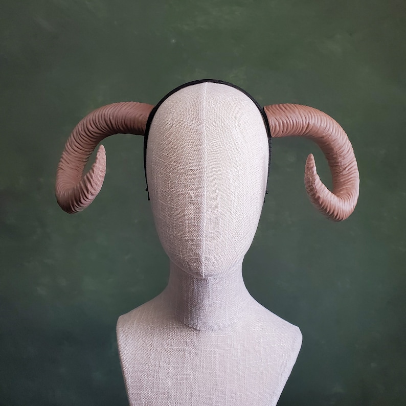 May include: A light brown headband with two curved horns. The headband is on a white mannequin head.