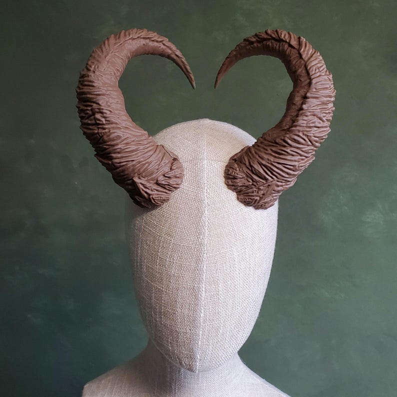 May include: A pair of brown, textured horns.  The horns are sculpted with a detailed, rough surface and curve gracefully outward. They are mounted on a light beige mannequin head, providing a clear view of the horns' shape and texture. These horns are ideal for costume creation, cosplay, or theatrical productions, adding a dramatic, fantasy element to any project.