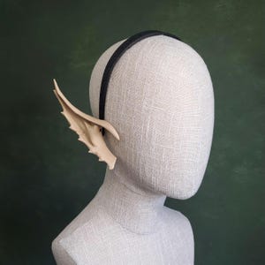 Puede incluir: Una diadema negra con orejas de elfo beige adjuntas. Las orejas son puntiagudas y tienen una apariencia texturizada en forma de ala. La cabeza del maniquí está sobre un fondo verde oscuro.