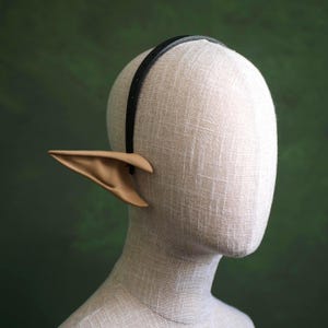 May include: A pair of tan elf ears attached to a black headband, displayed on a mannequin head. The ears are pointed and have a realistic appearance. The headband is thin and sits atop the head. The background is a solid, muted green.
