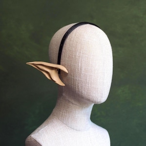 May include: A pair of prosthetic elf ears in a light beige color, attached to a black headband. The ears are detailed with realistic textures and are displayed on a mannequin head against a green backdrop.
