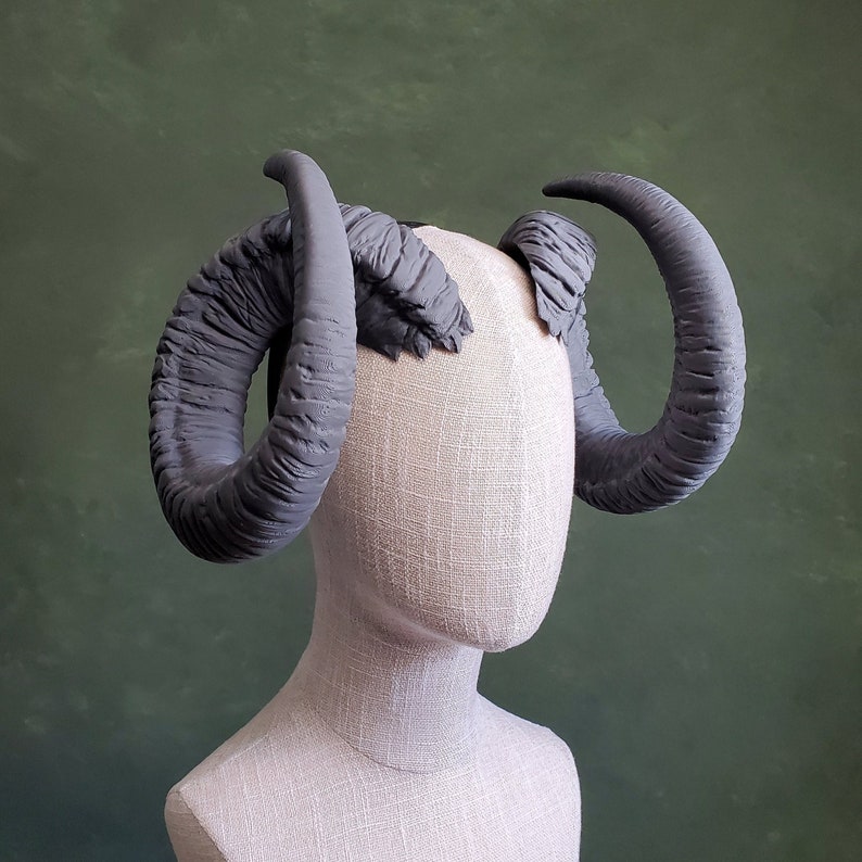 May include: A pair of gray, textured, sculpted horns on a white mannequin head. The horns are curved and pointed, and they are attached to the mannequin's head at the top of the forehead.