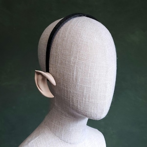 Puede incluir: Una diadema negra con orejas de elfo protésicas adjuntas. Las orejas son de un tono de piel pálido, con una forma curva y puntiaguda. La diadema es fina y oscura. La cabeza del maniquí está cubierta de tela clara.