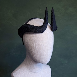 Puede incluir: Un tocado de cuernos negros con un acabado texturizado, exhibido en una cabeza de maniquí. Los cuernos son curvos y puntiagudos, con una apariencia oscura y mate. El tocado se asienta sobre una banda negra.