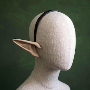 May include: A pair of elf ears in a light skin tone attached to a black headband. The ears are pointed and have detailed inner ear features. The headband is thin and sits atop a mannequin head against a green backdrop.