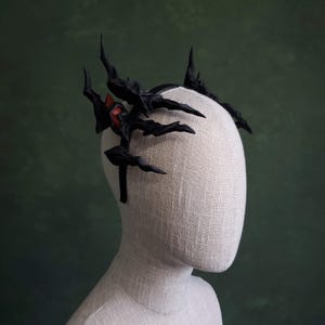 May include: A black headpiece with a spiky, ornate design, featuring sharp, angular elements and a touch of red. The headpiece is displayed on a mannequin head against a green backdrop, showcasing its gothic or fantasy aesthetic.