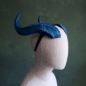 Puede incluir: Una diadema de cuernos azules sobre una cabeza de maniquí. Los cuernos son curvos y texturados, con un aspecto satinado. La diadema es negra y se asienta sobre la cabeza. El fondo es verde apagado.