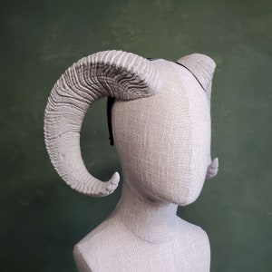 May include: A pair of white, textured, sculpted horns attached to a headband. The horns are curved and have a rough, organic texture.