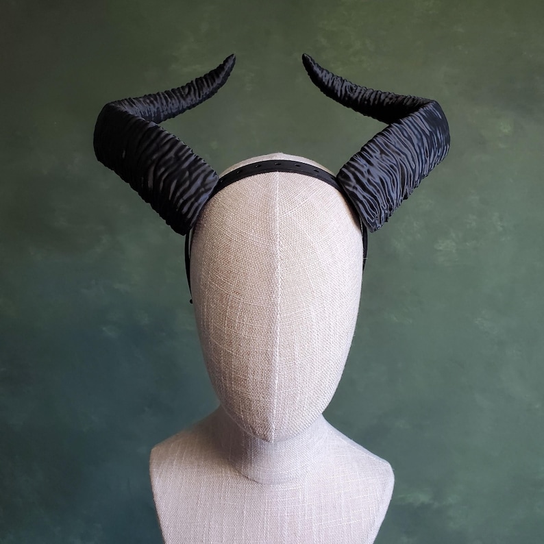 May include: Black devil horns headband.  The headband is made of a black, textured fabric. The horns are large and curve outward. The headband is shown on a mannequin head.  The horns are richly textured and have a slightly pleated appearance. The headband is a fashion accessory perfect for Halloween or costume parties.