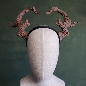 Puede incluir: Una diadema marrón con dos grandes cuernos de rama de árbol estilizados. La diadema está en una cabeza de maniquí blanca.