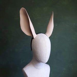 May include: A beige bunny ear headband with a black band, displayed on a mannequin head. The ears are long and upright, with a smooth texture. The background is a dark green.