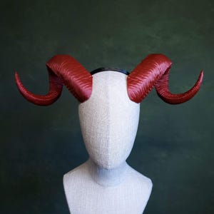 Puede incluir: Un par de cuernos rojos y curvados unidos a una diadema negra, exhibidos en una cabeza de maniquí. Los cuernos tienen una superficie texturizada y un color rojo intenso, para un disfraz o accesorio.