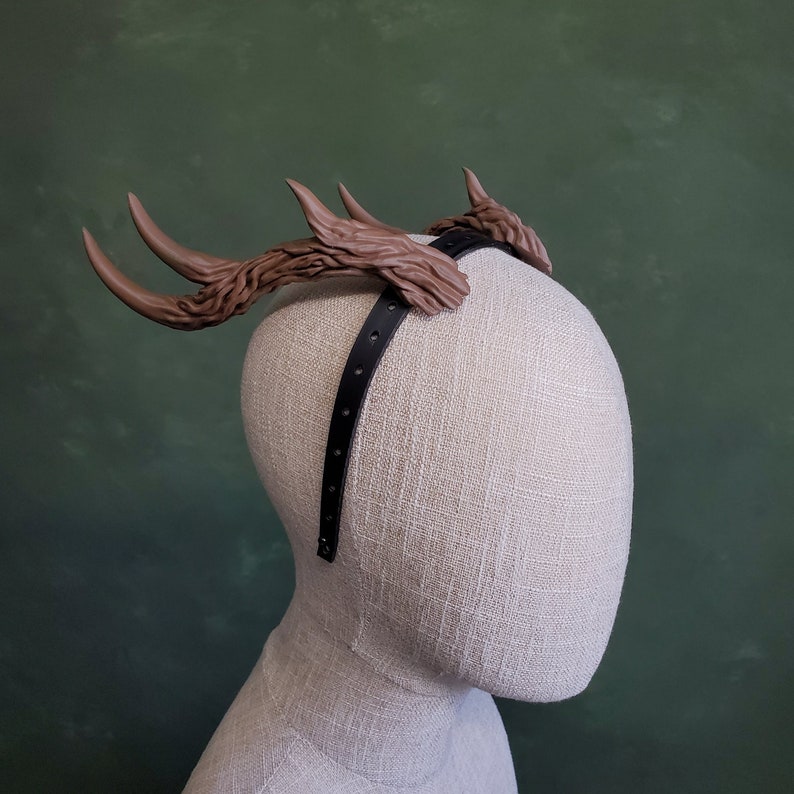 May include: A brown antler headband with a black elastic band. The headband is on a fabric mannequin head.