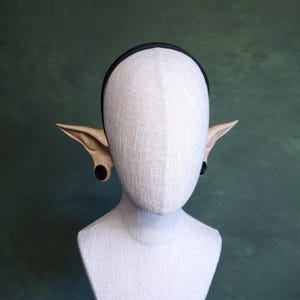 Puede incluir: Una cabeza de maniquí con una diadema negra y orejas de elfo adjuntas. Las orejas son de color beige claro con tapones negros. El maniquí está cubierto con una tela beige claro y el fondo es verde oscuro.