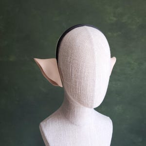 May include: A beige elf ear headband on a mannequin. The headband is black and the ears are a light beige color. The mannequin is white and the background is a dark green.
