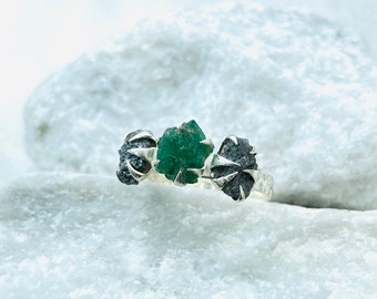 May include: A silver ring featuring three raw gemstones. The central stone is a green emerald, and the two flanking stones are black diamonds.