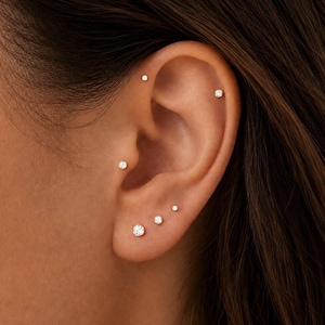 May include: A set of five small, round, clear stud earrings made of polished metal. The earrings are arranged in various positions along the earlobe and cartilage, featuring a simple, circular design with a single sparkling stone in each.