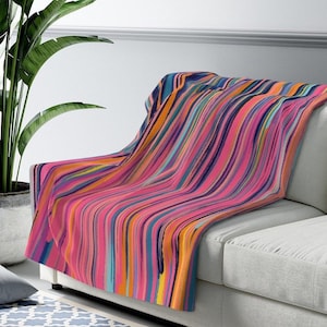 Può includere: Una coperta a righe colorata con sfondo rosa e righe verticali in tonalità di blu, arancione, giallo e viola. La coperta è drappeggiata su un divano grigio chiaro.