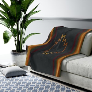 May include: A dark gray throw blanket with a geometric pattern in shades of orange, yellow, and green. The blanket is folded and draped over a light gray sofa.