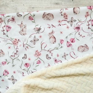 May include: A white baby blanket with a floral print featuring bunnies, butterflies, and birds. The blanket is reversible with a soft, cream-coloured fleece on the other side.