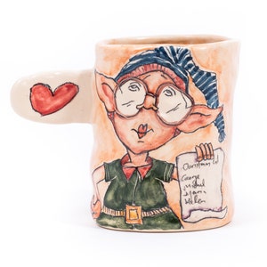 May include: A ceramic mug featuring a hand-painted design of an elf wearing glasses and a blue hat. The elf is holding a scroll that says "Christmas List" and has a heart-shaped handle.
