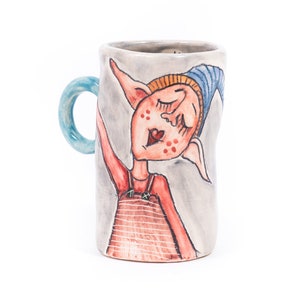 May include: A ceramic mug featuring a hand-painted design of a whimsical elf wearing a blue hat and red and orange clothing. The mug has a light blue handle.