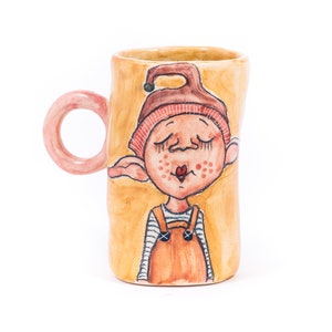 May include: A ceramic mug featuring a hand-painted illustration of a cartoon elf wearing a red hat and brown overalls. The mug is yellow with a pink handle.