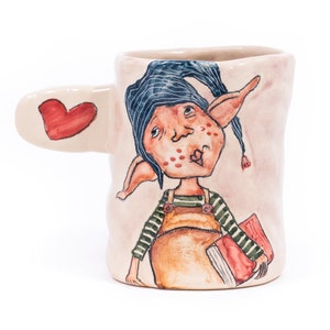 May include: A ceramic mug featuring a whimsical illustration of a cartoon elf wearing a blue hat, holding a book and with a heart-shaped handle. The mug is painted in shades of pink, blue, red, and yellow.