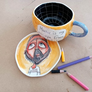 May include: A yellow ceramic mug with a black and white grid pattern inside. The mug has a blue handle and says "Music is my escape from..." on the side. A matching yellow ceramic saucer with a cartoon drawing of a person with a beard and a striped shirt is next to the mug. Three colored pencils are on the table.
