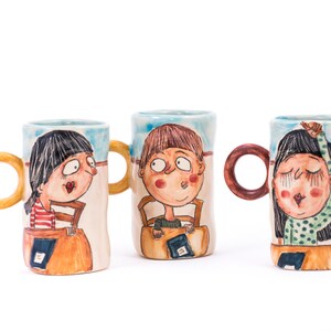 May include: Three ceramic mugs with hand-painted illustrations of children. Each mug features a different child with unique facial expressions and clothing. The mugs have colorful handles and are decorated with a blue and white background.