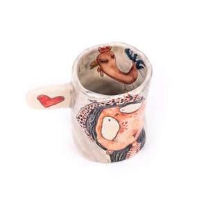 Puede incluir: Una taza de cer&aacute;mica con un dise&ntilde;o pintado a mano de una mujer con un coraz&oacute;n rojo en el asa. La mujer lleva un sombrero con estampado rojo y blanco y se puede ver un gallo dentro de la taza.