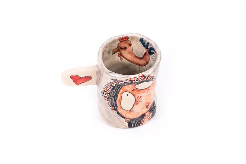 Puede incluir: Una taza de cer&aacute;mica con un dise&ntilde;o pintado a mano de una mujer con un coraz&oacute;n rojo en el asa. La mujer lleva un sombrero con estampado rojo y blanco y se puede ver un gallo dentro de la taza.