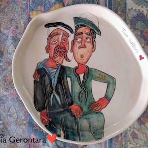Könnte beinhalten: Ein weißer Keramikteller mit einer handgemalten Illustration von zwei Comic-Matrosen. Die Matrosen tragen blaue und grüne Matrosenanzüge und haben rote und schwarze Gesichtszüge. Auf dem Teller steht der Schriftzug "Two Sailors".
