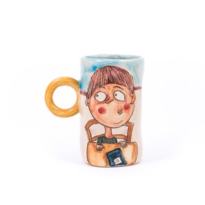May include: A ceramic mug with a hand-painted design of a person sitting in a chair and reading a book. The mug has a light blue background with a yellow handle.