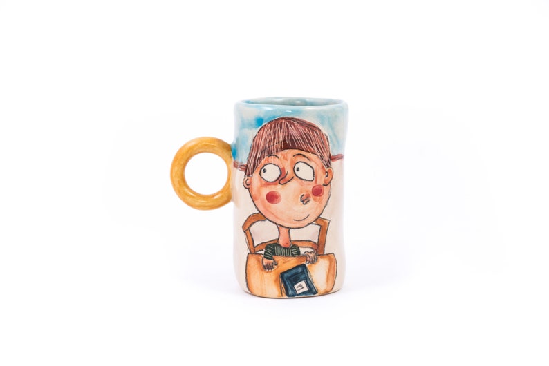 May include: A ceramic mug with a hand-painted design of a person sitting in a chair and reading a book. The mug has a light blue background with a yellow handle.